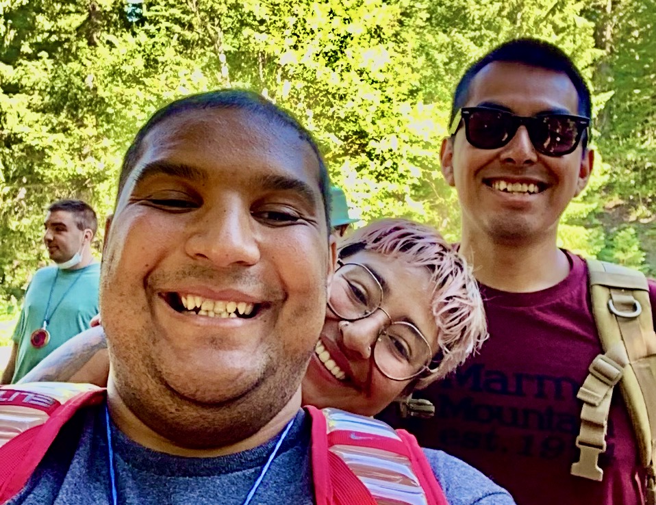 Smiling Camper in foreground, with two smiling counselors looking over his left shoulder. Sun shines on the evergreen trees in the background.