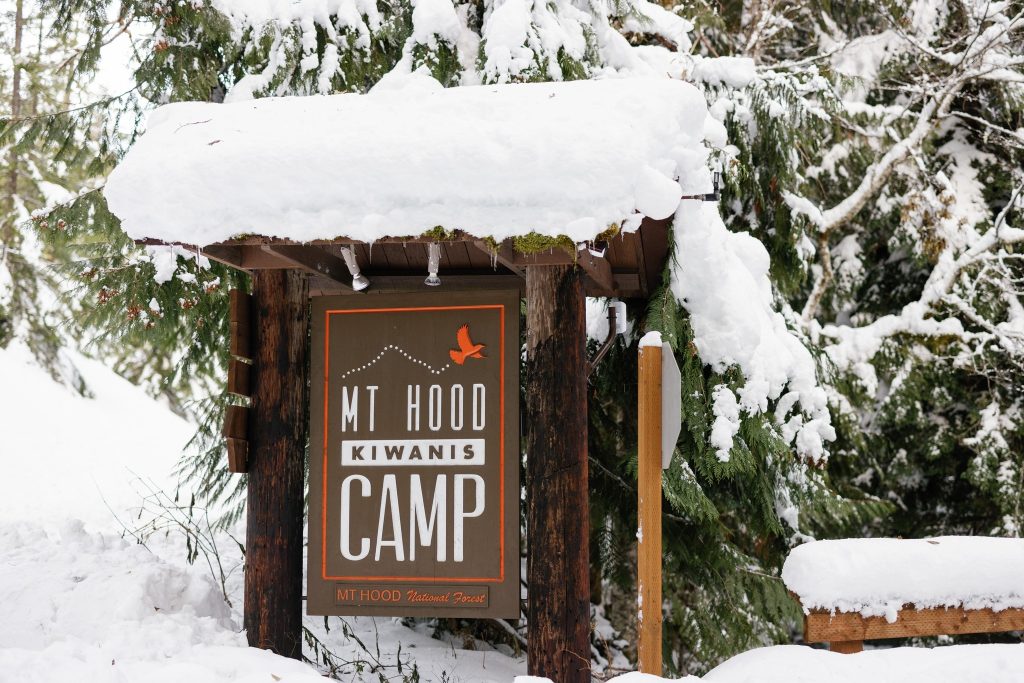 Wooden sign, covered in snow. Reads: Mt. Hood Kiwanis Camp: Mt. Hood National Forest.