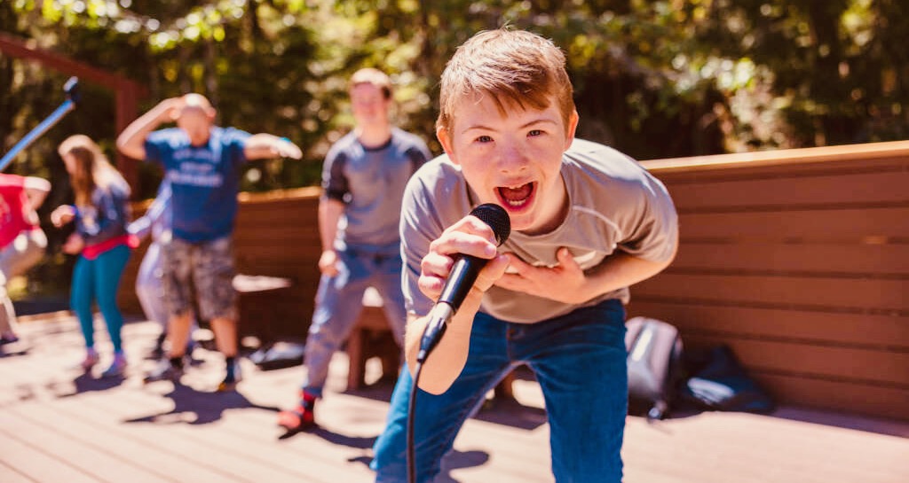 Camper on stage, singing into a microphone, leaning into the camera.