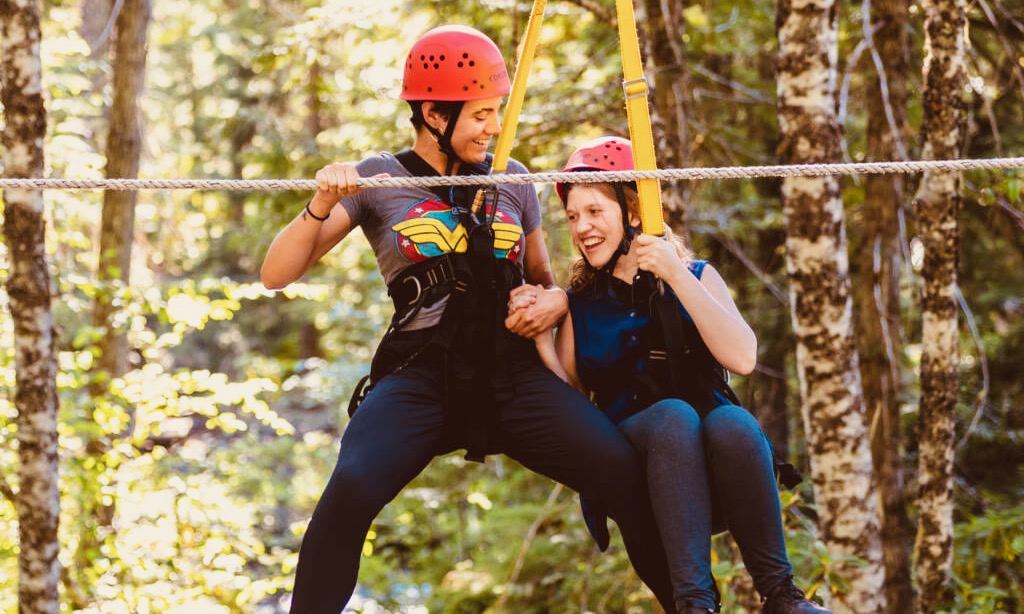 Camper and counselor in harnesses, crossing the stream hand-in-hand and smiling.