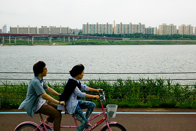 Two young people ride a tandem bicycle along a waterfront.