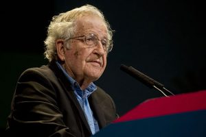 A photograph of Noam Chomsky sitting at a podium